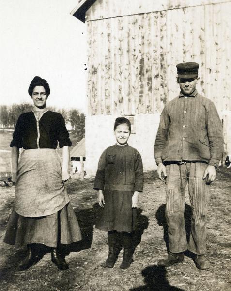 Photographic postcard of a farm couple and their daughter posing outdoors in front of a barn.