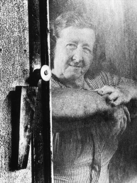 Woman looking out from behind a screen door, leaning on the door.