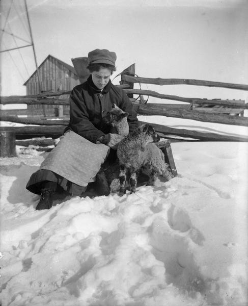 Winter scene with woman tending lambs in a snow-covered farmyard with log buildings in the background.