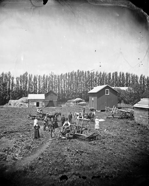 Family and farm implements in a field with farm buildings and haystacks in the background.