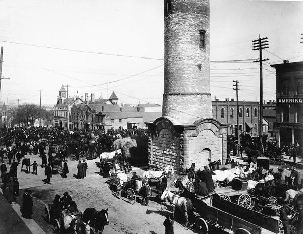 The monthly horse market on East Washington Avenue between Pinckney and Webster Streets. The 125-foot-high water tower (1890-1921) and Amerika, (1889-1922) office of the prominent Norwegian-American newspaper at the time, are also in view.