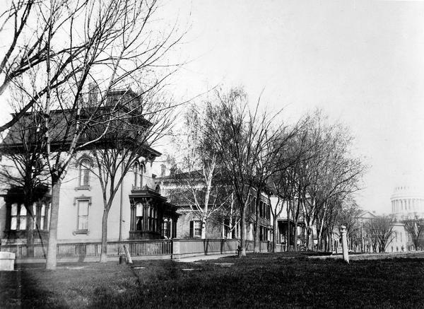 Several houses at the intersection of Wilson Street and Monona Avenue with the home of Simeon Mills, one of Madison's most prominent citizens, on the corner. Next to it is the Atwood/Buck-Neckerman house. The Wisconsin State Capitol is in the background.