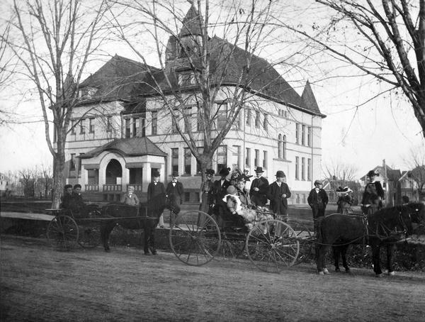 Exterior of the 6th Ward School, 1237 Williamson Street, built in 1894. Several people are posing on the road and sidewalk in front of the building with two horse-drawn buggies. The building was designed by the Madison firm of Conover and Porter, and featured five turrets, the tallest of which stood nine stories above the ground. It was finished with Ashland brown stone and cream brick. Renamed Marquette School in 1904. Torn down in April 1952.