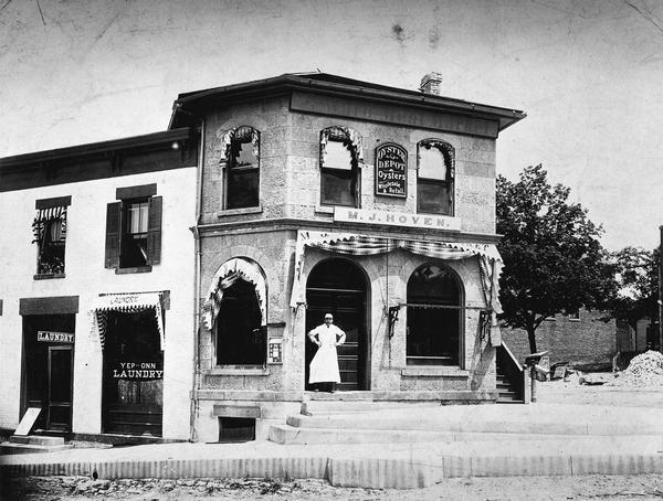 Matthew J. Hoven with hands on hips standing in front of M.J. Hoven's Meat Market. A sign on the building reads: "Oyster Depot; Wholesale & Retail". There is a rack on the right front window on which meats were hung for shoppers to inspect.  The Hoven building is at the corner of North Hamilton and East Mifflin Streets, which is now occupied by the Blue Marlin restaurant.