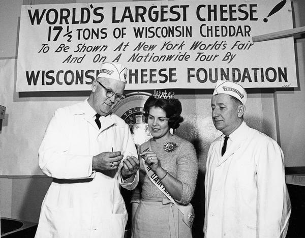 Alice in Dairyland, and two employees of Steve's Cheese, sample a piece of the World's largest cheese. The gigantic piece of cheese was part of Wisconsin's exhibit at the 1964 World's Fair held in New York.