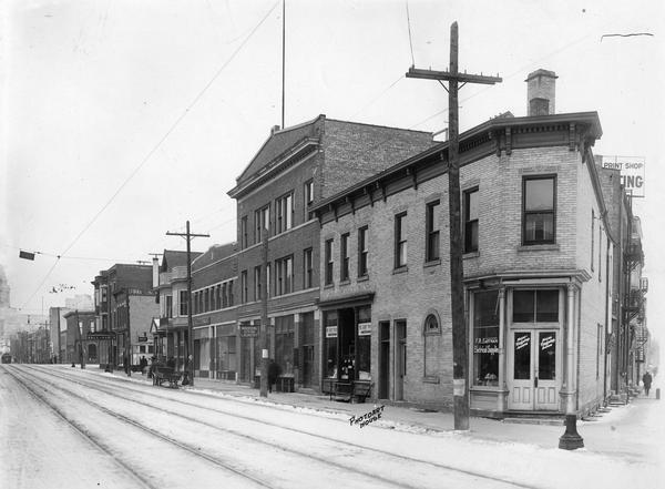 View of the 400 block of State Street, south side with the following businesses: 437 Fred R. Eastman Electrical Supplies, 435 M.J. Levin's The Square Tailors, 433 Wisconsin School of Music, 429 madison Steam Laundry, 421 Capitol City Meat Market, 419 Grocery, 417 Elizabeth Taggart's Kopper Kettle Tea Shop, 415 Ralph Richardson's Lunch Wagon, 413 Klein-Statz Wallpaper, and the Wisconsin State Capitol in the background.
