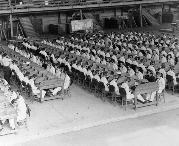 Navy personnel sitting at typewriters wearing headphone learning code, probably during World War II.  The sailors are in an auditorium or gymnasium, possibly the Field House on the University of Wisconsin-Madison, campus.