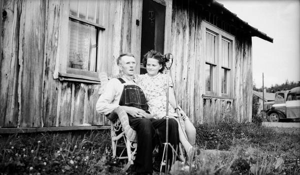 Charlie Spencer (b.1873?), a Kentuckian who migrated to Crandon, recorded white spirituals for song collector Helene Stratman-Thomas. He is sitting in a chair outdoors in front of a building. A young girl is sitting in his lap, and there is a microphone in front of them.