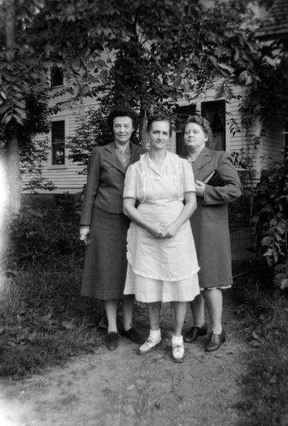 A group portrait of three women. (l to r) Song collector Helene Stratman-Thomas, Mrs. Kamma Grumstrup, a singer of old ballads in Danish, and possibly Berit Sanford. They are standing on a dirt path through a yard with a house and trees in the background.