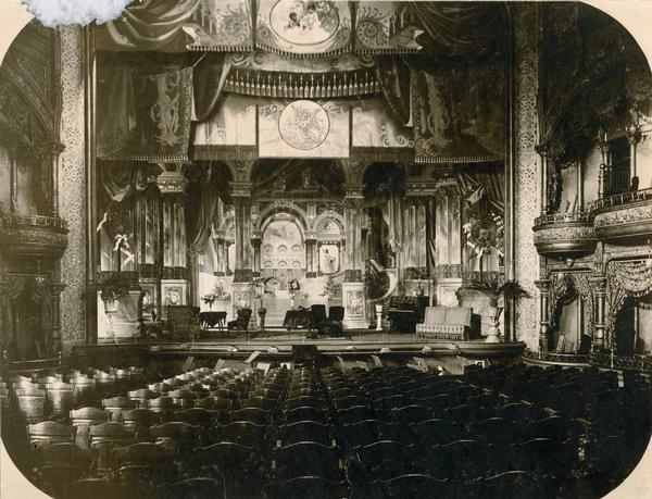 Interior of the Fuller Opera House looking over seats to the stage with 1,200 seats on a main floor, a second-level balcony, a third-level gallery, and ten private boxes.  There was a separate foyer and stair tower to the gallery.  Interior features include parquet floors, gold-gilt and amber walls, and 500 gas and electric lights.