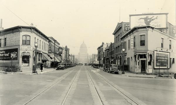 View of State Street with the Wisconsin State Capitol in the background. Included in the view is Dan's Shoe Repairing, New Place Cafe, Huegel & Hyland Shoes, Treasure Trove, Chop Suey Restaurant, the Wisconsin Lunch and other shops. There are automobiles parked curbside, streetcar tracks and billboards for Chesterfield cigarettes, and a Dodge Brothers Sedan.