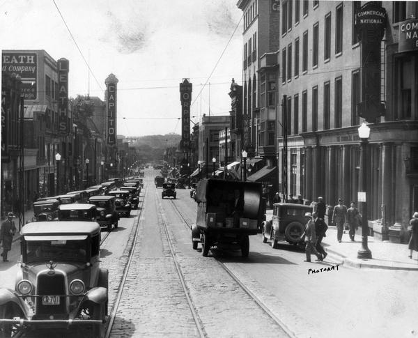Slightly elevated view of heavy automobile traffic, delivery trucks and pedestrians on State Street at North Carroll Street. View reaches to Bascom Hill of the University of Wisconsin. Commercial buildings with Leath's, Capitol Theatre, Orpheum Theatre, YWCA, Commercial National Bank and other shops are on both sides of State Street.