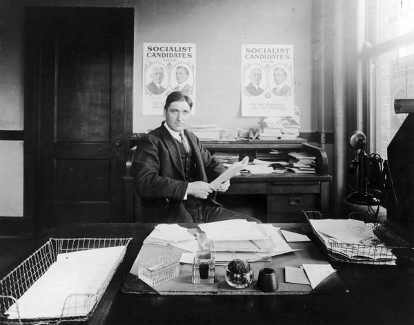 Adolf Germer seated in his office with posters for socialist candidates on the wall behind him.