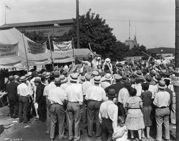 Slightly elevated view of a large crowd gathered around two men handing out souvenir International Harvester yardsticks at the Indiana State Fair.
