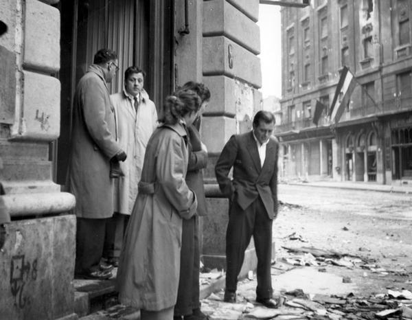 Russell Jones, facing camera wearing a light trench coat, standing with a group of journalists on a street corner during the fighting in Budapest, Hungary. After most reporters had left the city, Jones stayed in place during the worst of the fighting after Soviet troops invaded. Russell Jones won a Pulitzer Prize for his reporting about the revolt.