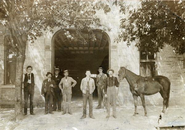 View from street of John Hess and Froderick Schmitz and a horse on the sidewalk in front of the entrance to 508 State Street, the Hess and Schmitz Livery Boarding and Sale Stable. There are five unidentified men standing nearby.