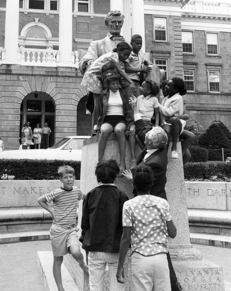 Governor Warren P. Knowles reaches up to touch the hand of a girl at the Lincoln Memorial on the University of Wisconsin-Madison campus. The girl seated, center, is Mary Wells and her brother Eric Wells is seated above her. Tammy Todd (top left bending over to touch Gov. hand, and Tina Wells seated on left.