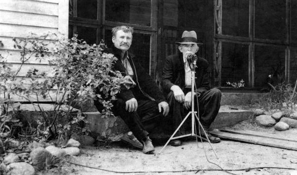 Charles Robinson, singer of lumberjack songs, left, with an unidentified man sitting behind what appears to be a microphone on a stand.