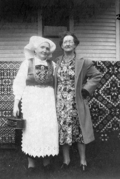 Mrs. Brithe Lothe, age 75, and Mrs. Hannah Haug, singers of Norwegian folk songs. Behind them are two quilts or rugs hanging over what may be porch railings of a house.