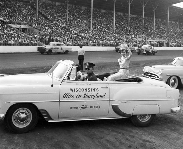 Mary Ellen Jenks, the 1953 Alice in Dairyland from Chippewa Falls, rides in the official convertible provided for Alice by Hult's in Madison. She is waving to the crowds gathered in the grandstand to watch stock car races at the Wisconsin State Fair. A uniformed man drives Alice's car, and several of the race cars are seen in the background.