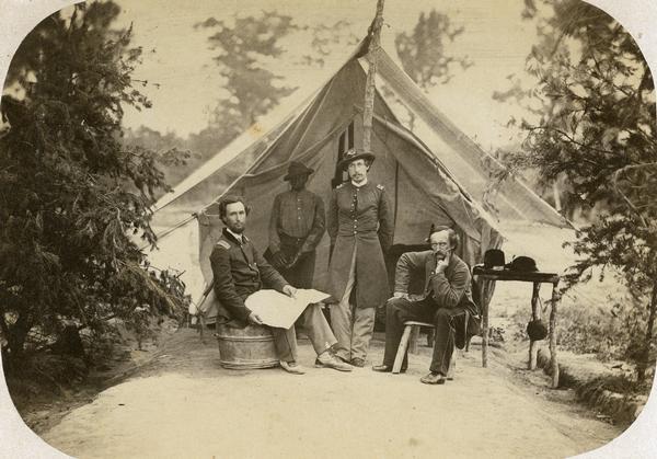 Officers of Company I, 7th Wisconsin Infantry: (left to right) Lt. Joseph N.P. Bird, Captain George H. Walther, and Lt. Christopher Lefler. Their Black servant, Alonzo Gambel, can be seen standing in the shadow of the tent. This photograph was taken during the summer of 1862 while the regiment was camped near Fredericksburg, Virginia.