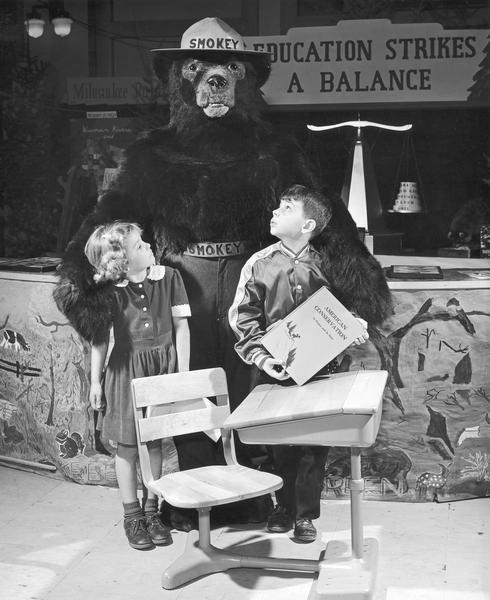 Man in a Smokey The Bear costume poses behind a school desk with a boy and a girl.