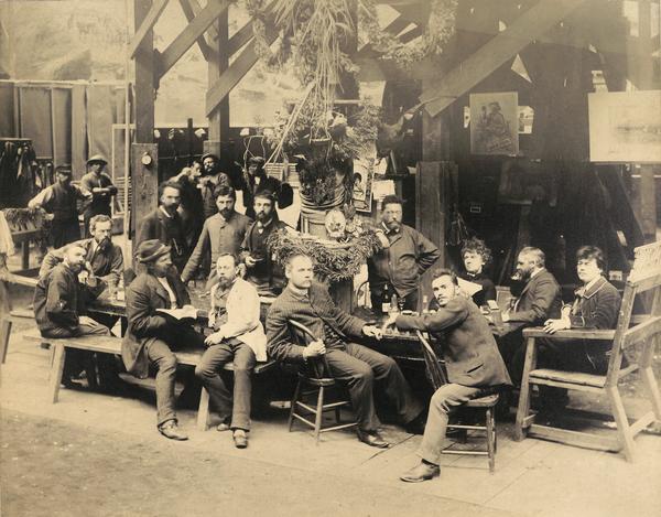 Group portrait of German painters relaxing in the studio of the American Panorama Company, during a break from painting the Jerusalem cyclorama depicting the crucifixion of Christ. Artists with their specialties include from the left, standing at the table: Franz Bilberstein (landscapes), Richard Lorenz (animals), Johannes Schulz (figures), and Bernhard Schneider (landscapes); and sitting from the left end of the table: Bernhard (Wilhelm?) Schroeder (Schroeter) with pipe (landscapes), Franz Rohrbeck (figures, especially Confederate), Friedrich Wilhelm Heine (wearing a hat) (Supervisor and master of composition), Karl Frosch (Frosh), Thaddeus Zukotynski (Zuchatinsky) (figures), George Peter (animals), Amy(?) Boos (Boss), August Lohr (in profile) (Supervisor and designer of landscape settings), and Herman Michalowski (figures).