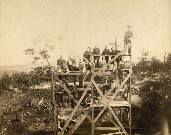 Group portrait of a group of German panorama painters on a scaffolding in their Milwaukee studio, with The Atlanta Cyclorama as a backdrop. Included in the group are, standing from the top left: Franz Bilberstein (landscapes), August Lohr (supervisor and designer of landscape settings), Herman Michalowski (figures), Feodor von Luerzer (landscapes), Franz Rohrbeck with flag (figures, especially Confederates), Theodor Breidwiser (figures), Johannes Schulz (seated) (figures), and Otto Dinger at the top right (figures). In front of them from the left with large moustache, Albert Richter (figures), Gustav Wendling (figures), Bernhard Schneider (landscapes), Bernhard (Wilhelm?) Schroeder (Schroeter?) (landscapes), and Paul Wilhelmi (figures). Standing in the center are Friedrich Wilhelm Heine (supervisor and master of composition) with Theodore R. Davis, a reporter who had reported on the battle. Robert(?) Schade is one of the men also seated at that level.