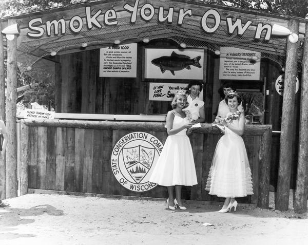 1952 Alice in Dairyland Beverly Ann Steffen and 1953 Alice in Dairyland Mary Ellen Jenks standing in front of a "Smoke Your Own Fish" stand at the 1953 Wisconsin State Fair. The stand was sponsored by the State Conservation Commission of Wisconsin to promote the eating of carp.