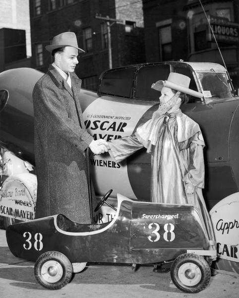 Meinhardt Raabe, dressed as the Coroner from "The Wizard Of Oz," shaking hands outdoors with a man wearing a hat and coat. Behind them is the Oscar Mayer Wienermobile, and in front of the men is a Speed-o-Racer, a small toy car that has a sign painted on it that reads: "Supercharged 38."