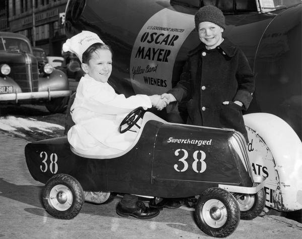 Meinhardt Raabe, dressed as Little Oscar, is sitting in a Speed-o-Racer and shaking hands with Billy Marble. Behind them is the Wienermobile.