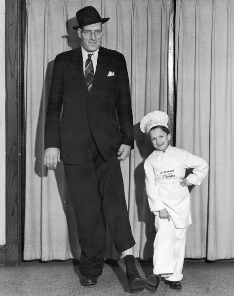 A giant man, wearing a suit and hat, and Meinhardt Raabe, who is dressed as Little Oscar, are standing together and pulling up their pants legs to compare shoe sizes.