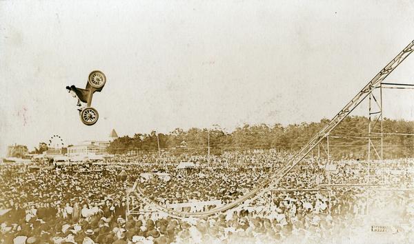 A female daredevil stunt driver, possibly Beryle Elliot, in a horseless carriage catapults up into the air, doing a loop-the-loop or sommersault off a ski jump-type apparatus at the Brockton, Massachussetts Fair where a huge crowd has assembled.