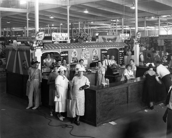 Elevated view of employees and patrons gathered around a beer and sandwich concession stand on the midway at the Wisconsin State Fair, where Gettelman On Tap is sold for 5 cents a glass, and a sign on the wall proclaims "This is a Union Bar!"