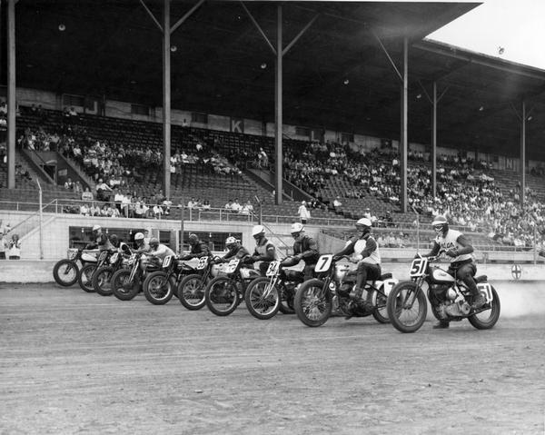 Ten motorcycle racers line up abreast on the track at the Wisconsin State Fair Park. A group of spectators watches from the stands.