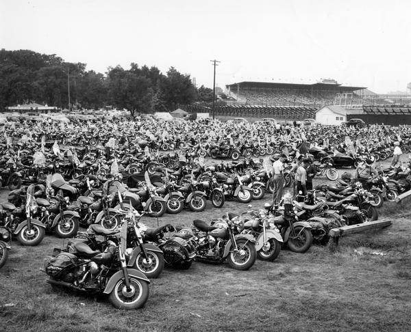 Hundreds of motorcycles are parked side by side in rows in a Wisconsin State Fair parking lot.  The grandstand and spectators appear in the background of the photograph.