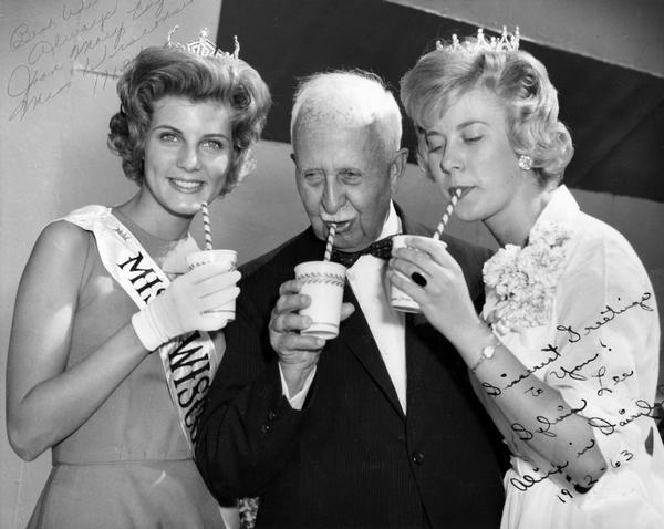 Miss Wisconsin 1962, Joan Mary Engh, and Alice in Dairyland 1962-63, Sylvia Lee, enjoy a drink of milk with an elderly gentleman during their visit to the Wisconsin State Fair. The print is autographed by both women.