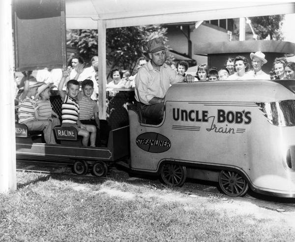 Boys wave from the passenger cars of Uncle Bob's train, while a crowd watches the amusement park ride at the Wisconsin State Fair.