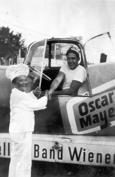 Meinhardt Raabe, dressed as Little Oscar and wearing a hat that reads "Yellow Band Wieners," is standing in front of the Wienermmobile and holding one side of the driver's side window open, while shaking hands with a man wearing an Oscar Mayer hat who is sitting behind the steering wheel.