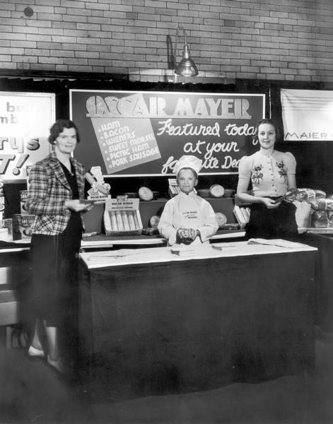 Meinhardt Raabe, dressed as Little Oscar, is standing behind an Oscar Mayer display counter between two women.