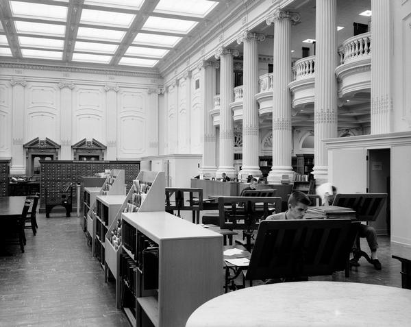 A view of the Library Reading Room in 1955, after the modernization of the 1950s was complete.  Most prominent is the division of the room into functional areas:  bound newspaper were read on stands in the area to the right; the west wall was lined with cubicles for microfilm reading; a new centralized circulation/reference desk stood has been moved into the room near the columns; and behind the card catalog were only a few of the original mahogany tables. Out of the frame to the left was a reading area with comfortable chairs. Other changes included modern furniture and acoustical tile and fluorescent lighting in the ceiling.