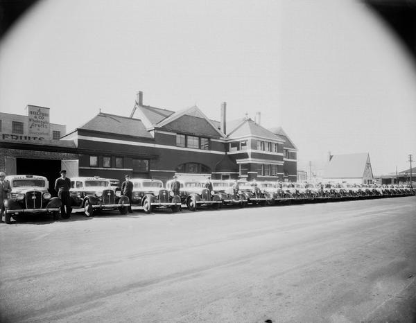 Lineup of 24 Ford City Car Company taxicabs and their drivers in front of City Car Co. office, located at 601 - 603 West Washington Avenue, (former Illinois Central Railroad Station) and Fiore Service Station, 601 West Washington Avenue. The view also shows J. Heilprin & Co. wholesale fruits and vegetables sign on side of their building, 612 W. Main Street.