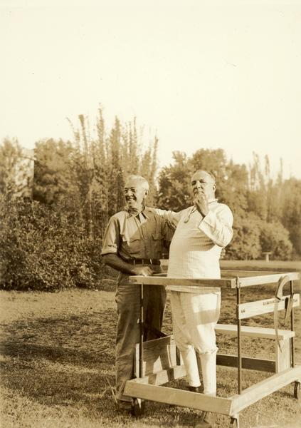 Raymond Robins and a bespectacled man standing together outdoors. Robins is standing inside a wooden frame on wheels.