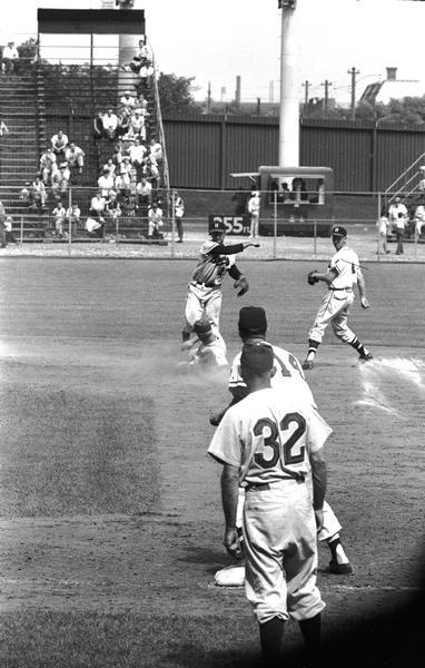 Milwaukee Braves shortstop Johnny Logan jumps over a sliding baserunner and throws to Braves' first baseman Frank Torre in a game at County Stadium. Second baseman Red Schoendienst looks on from the right. Schoendienst's first home game as a Brave after being acquired from the New York Giants was June 17, 1957.