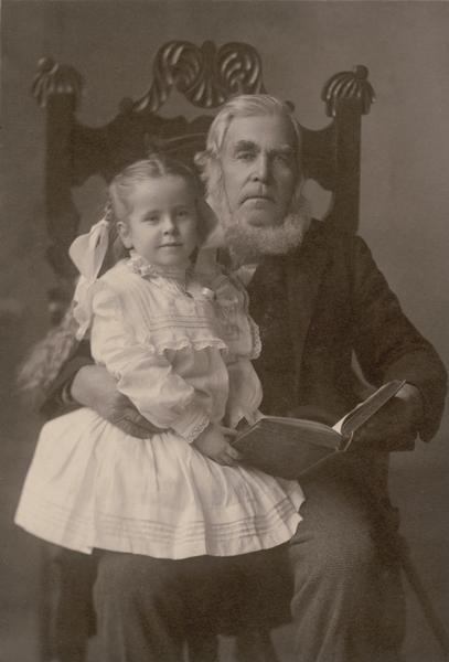 Studio portrait of Lars Davidson Reque, age 90, seated showing a book to his grandniece Martha Rothie Nelson, age 5.