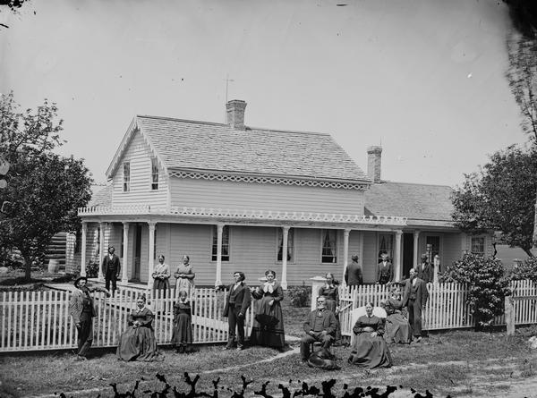 Family and frame house of Lars David Reque. The family is posing in the yard which is surrounded by a picket fence, with a large frame house behind with carpenter's lace on porch and festoon trim on roof; there is a wood barrel in the background on the left. A dog is sitting on the grass at the feet of a man in the foreground. Identified from photograph owned by Mary Rauschenberger, Madison, through Mike Bovre. One man is posing with his back to the camera.