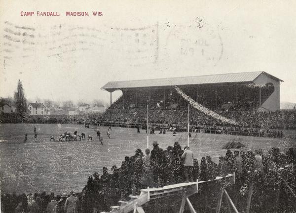 Elevated view of a football game at Camp Randall Stadium at the University of Wisconsin-Madison. Spectators are standing and sitting in the stands in the foreground. Caption reads: "Camp Randall, Madison, Wis."