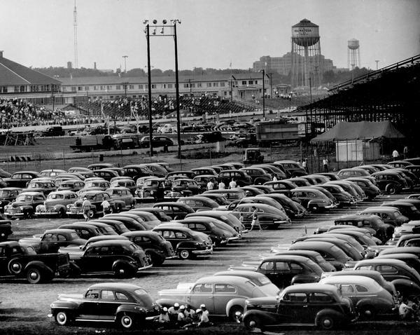 Left side of an elevated panoramic view of a full parking lot at the Wisconsin State Fair, with the grandstand and water towers in the background.