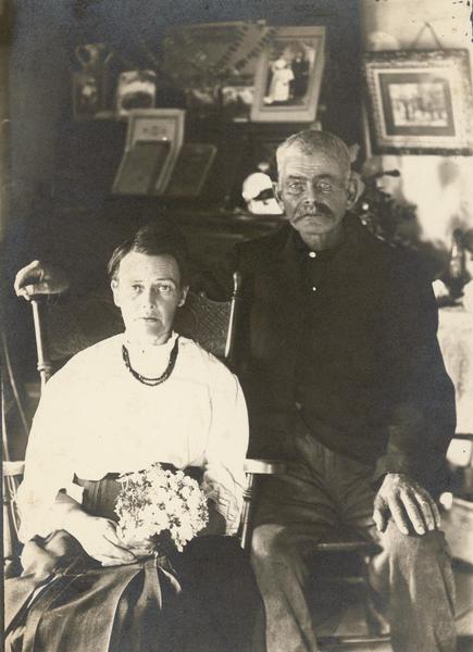Portrait of an elderly couple seated in their home. The woman is holding a bouquet of flowers.  The man is seated slightly behind her, with his right arm over the back of her chair, the other on his knee.