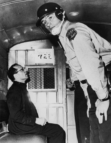 Father Groppi is seated in the back of a police wagon with police officer after being arrested in front of his parish for the second night in a row.